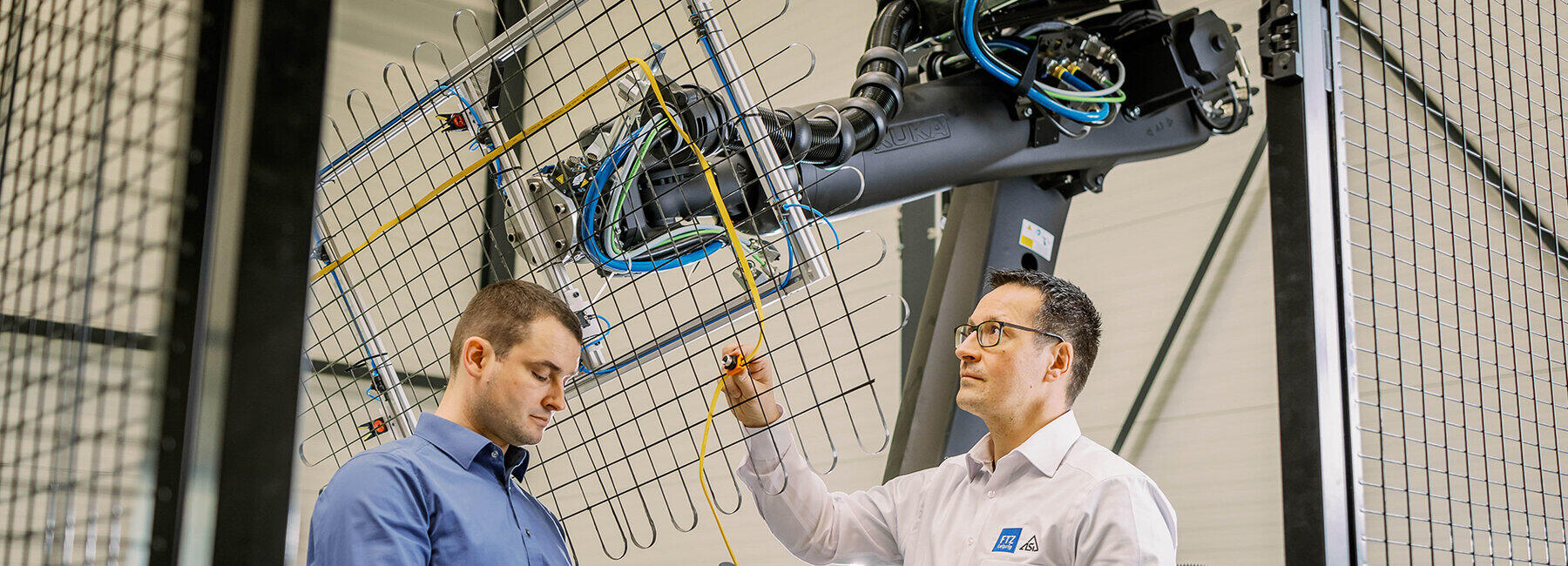 Zwei männliche Personen stehen vor einem Gitterbauteil, welches von einem Roboterarm gehalten wird