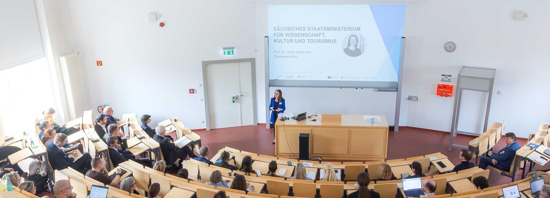 A perspective view of a lecture theatre; some seats are occupied; a woman in a blue blazer is standing at the lectern