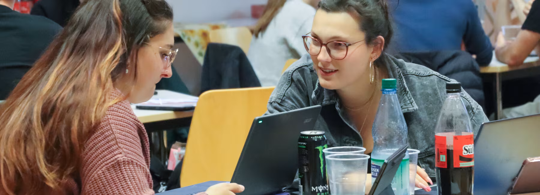 Zwei junge weibliche Personen an einem Tisch mit Laptops, Bechern, Colaflaschen und Energydrinks in Dosen. Die Frau rechts wendet sich der linken interessiert zu. Im Hintergrund sind weitere Personen an Tischen zu sehen.