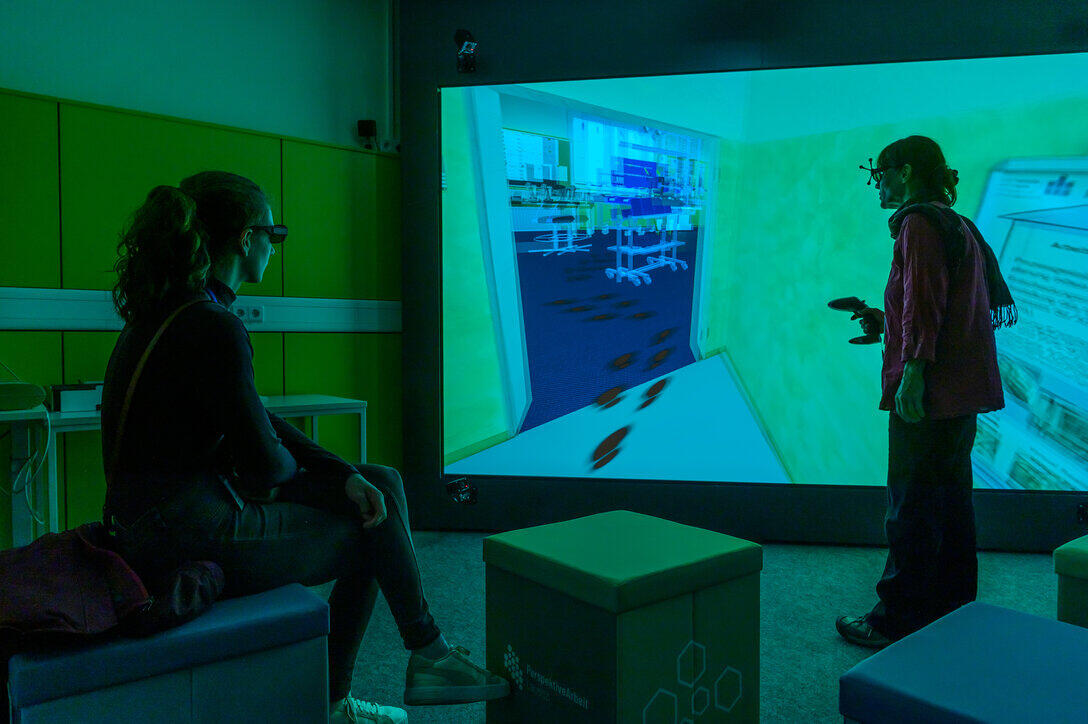 vor einer VR-Wand steht eine Person und eine Weitere sitzt etwas im Hintergrund