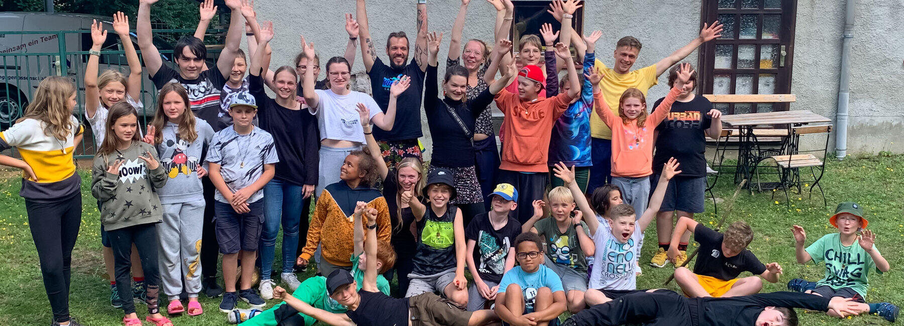 Gruppenbild auf einer Wiese vor einem Gebäude mit rund 30 Personen, darunter überwiegend Kinder zwischen 8 und 12 Jahren. Die meisten stehen jubelnd mit nach oben gestreckten Armen, andere sitzen auf dem Boden, einige liegen.
