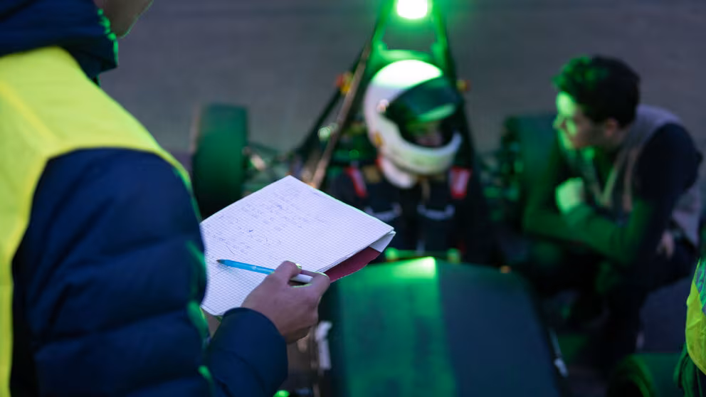 Im Bildvordergrund ist links im Anschnitt und von hinten eine Person mit Warnweste und einem Schreibblock zu sehen. Im Bildhintergrund sind unscharf der Fahrer mit Helm im Rennwagen und daneben eine knieende männliche Person im Gespräch miteinander zu sehen.
