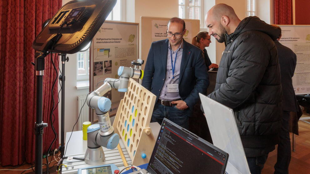 Zwei männliche personen stehen vor einer 4-gewinnt-Wand mit einem Roboterarm
