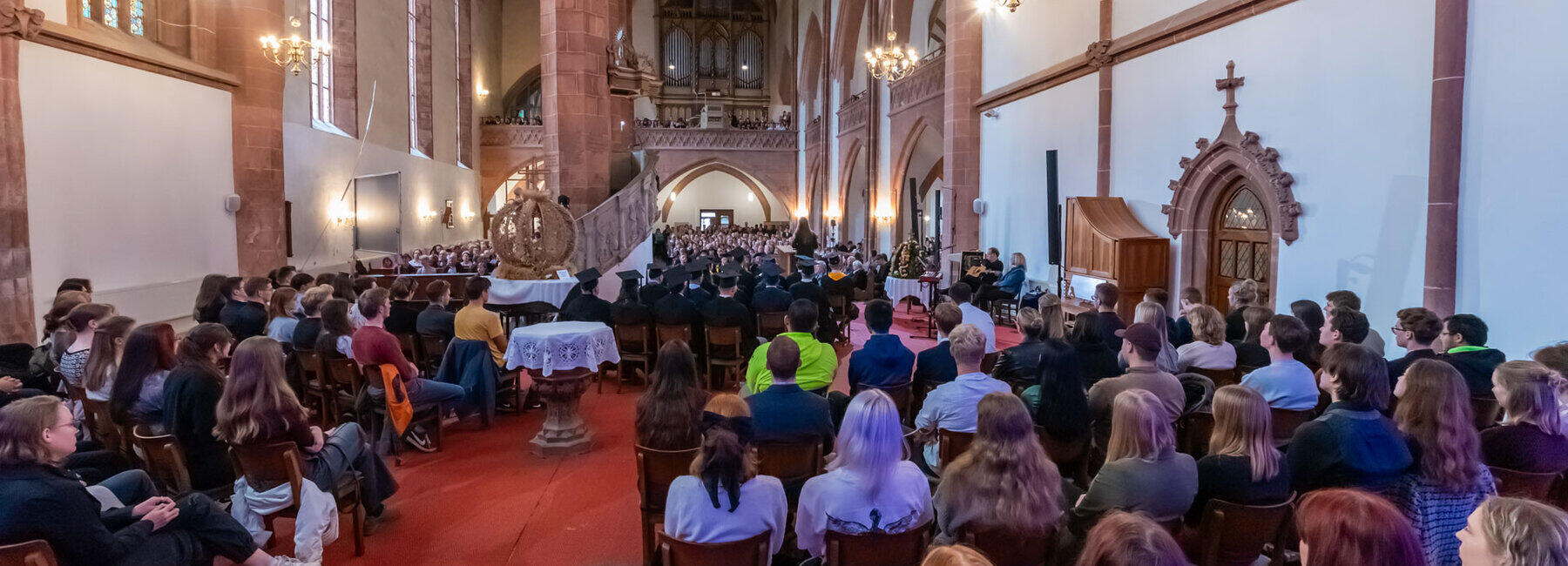 Blick vom Altarraum ins vollbesetzte Kirchenschiff aus dem Altarraum. Zwischen den Köpfen junger Menschen und den Doktorhut-geschmückten Köpfen von Professor:innen steht eine große Erntekrone.