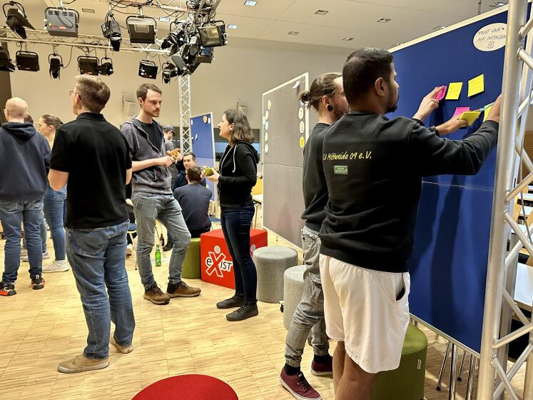 Gruppe von Personen steht im Raum und vor einer Pinnwand