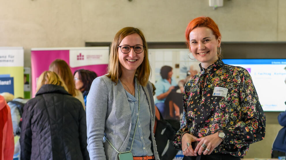 Zwei jüngere Frauen stehen in einem Foyer nebeneinander und schauen strahlend in die Kamera. Im Hintergrund sind unscharf weitere Menschen vor Rollups zu sehen. Auf einem der Rollups ist das Logo der Hochschule Mittweida zu erkennen.