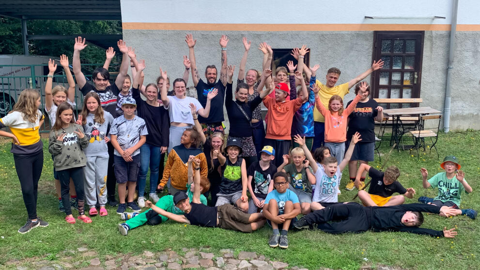 Gruppenbild auf einer Wiese vor einem Gebäude mit rund 30 Personen, darunter überwiegend Kinder zwischen 8 und 12 Jahren. Die meisten stehen jubelnd mit nach oben gestreckten Armen, andere sitzen auf dem Boden, einige liegen.