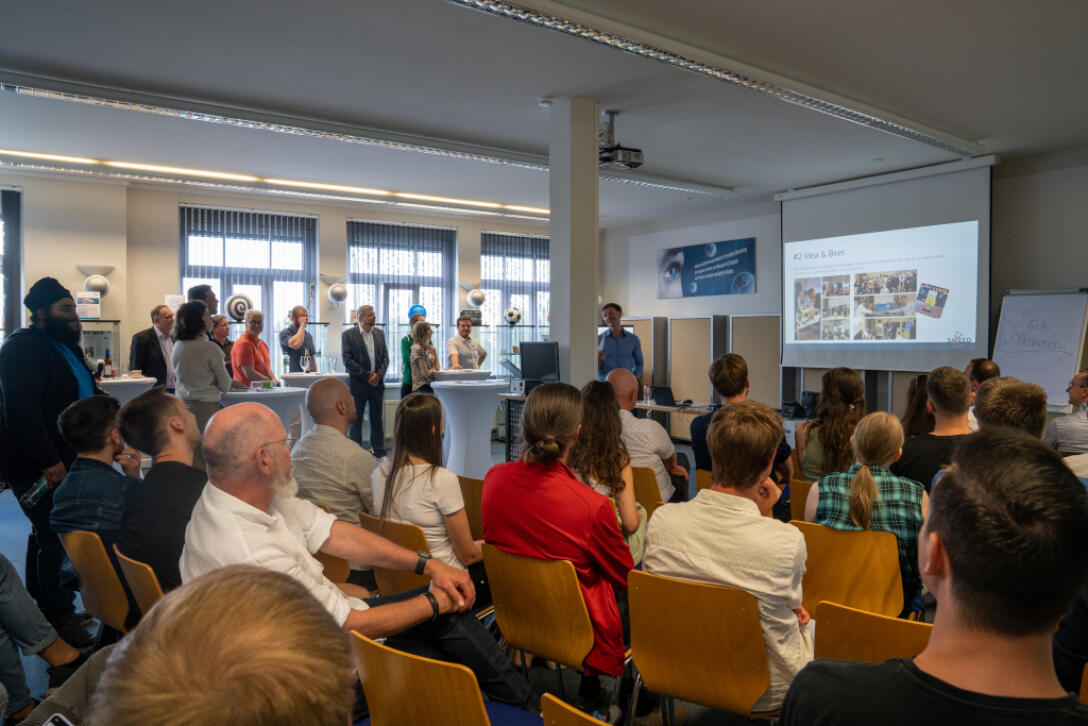 Besucher sitzen in Reihen und einige stehen an Stehtischen; Moderator steht an der Leinwand und präsentiert