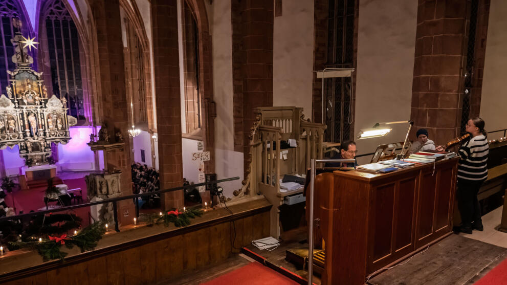 Blick auf Empore und Kirchenschiff. Auf der Empore sitzt eine männliche Person am Spieltisch der Orgel, rechts daneben stehen zwei weibliche Personen, eine spielt Geige, die andere Querflöte.