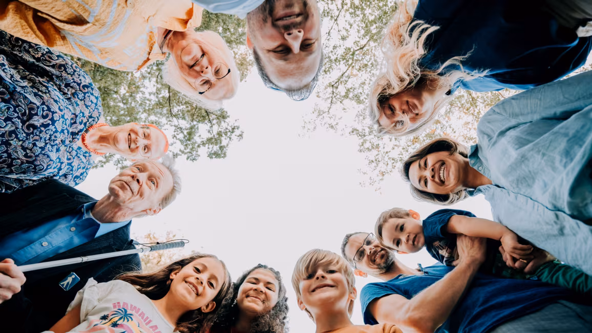 Eleven people of different ages, genders and skin colours are standing in a circle, looking down at the camera. Above them, in the centre, there is an unobstructed view of the sky.