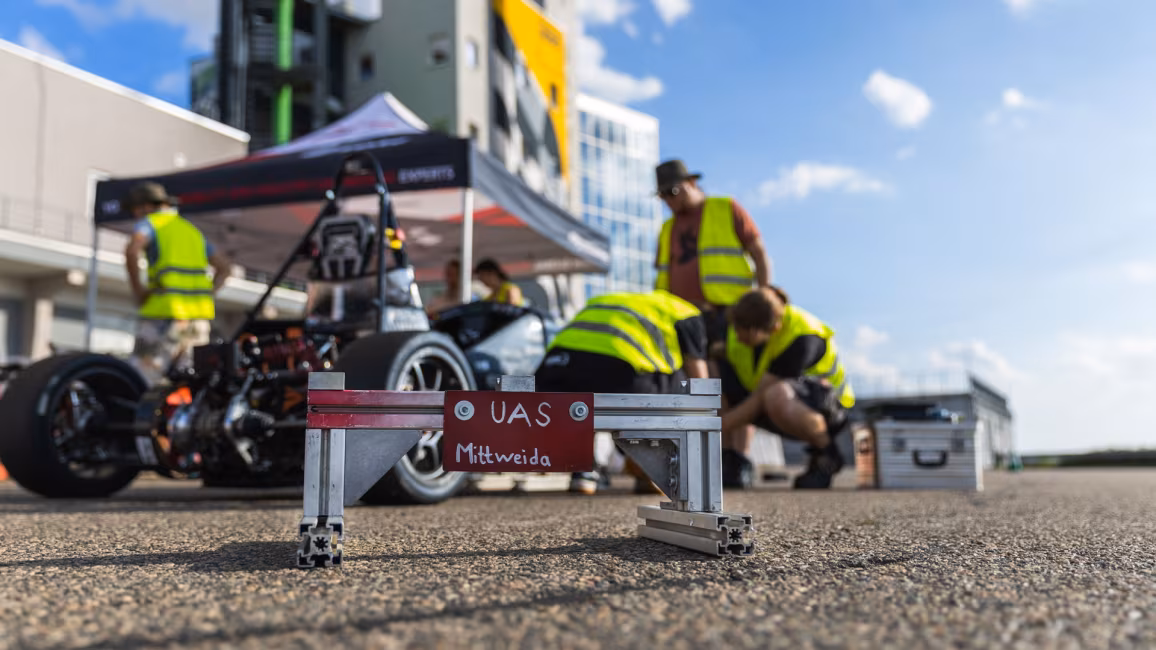 Ein Schild mit dem handgeschriebenen Text „UAS Mittweida“ steht auf dem Boden vor einem Rennwagen, an dem Personen hantieren. Im Hintergrund ist unscharf ein hohes Gebäude vor blauem Himmel zu sehen.